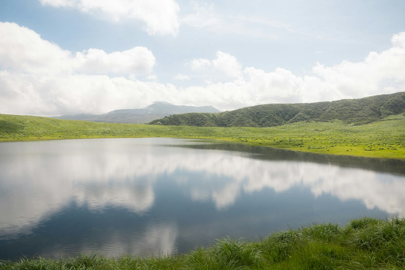 Calm lake reflecting green hills and clouds under a blue sky — still waters and green pastures