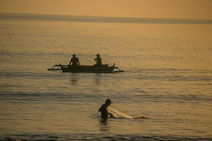 A person casting a fishing net in shallow water near a boat